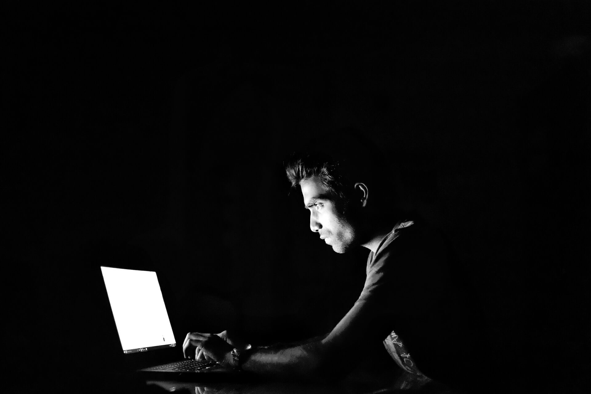 Men is sitting in front of the open laptop in the dark room